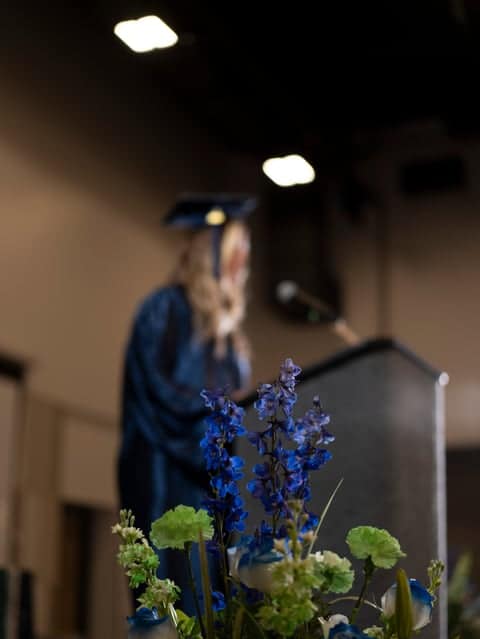 High School Graduate in a blue cap and gown, speaking through a mic with flowers in the forground.